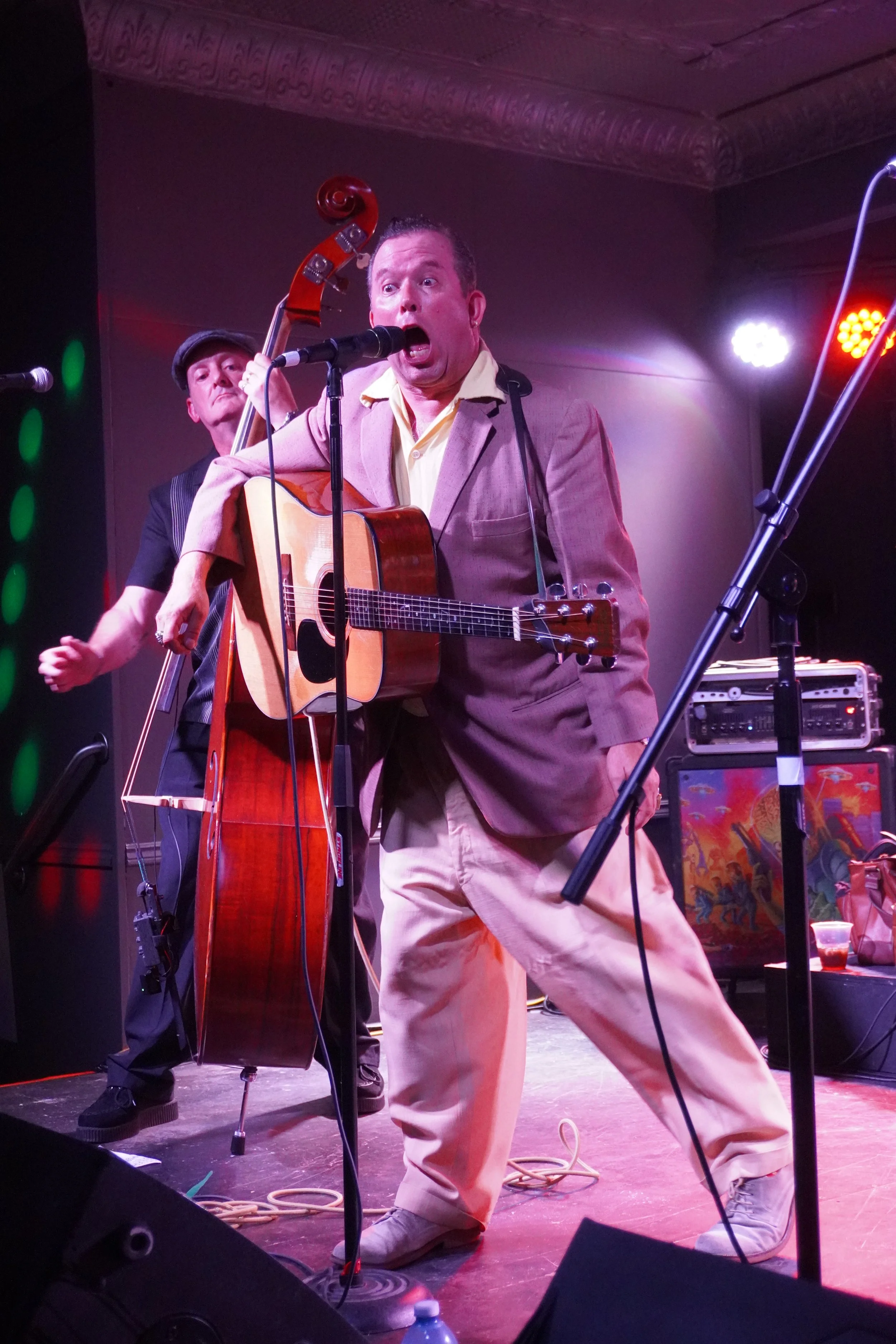 Man singing passionately into a microphone while playing an acoustic guitar on stage, with another musician playing a double bass in the background, during a live performance.