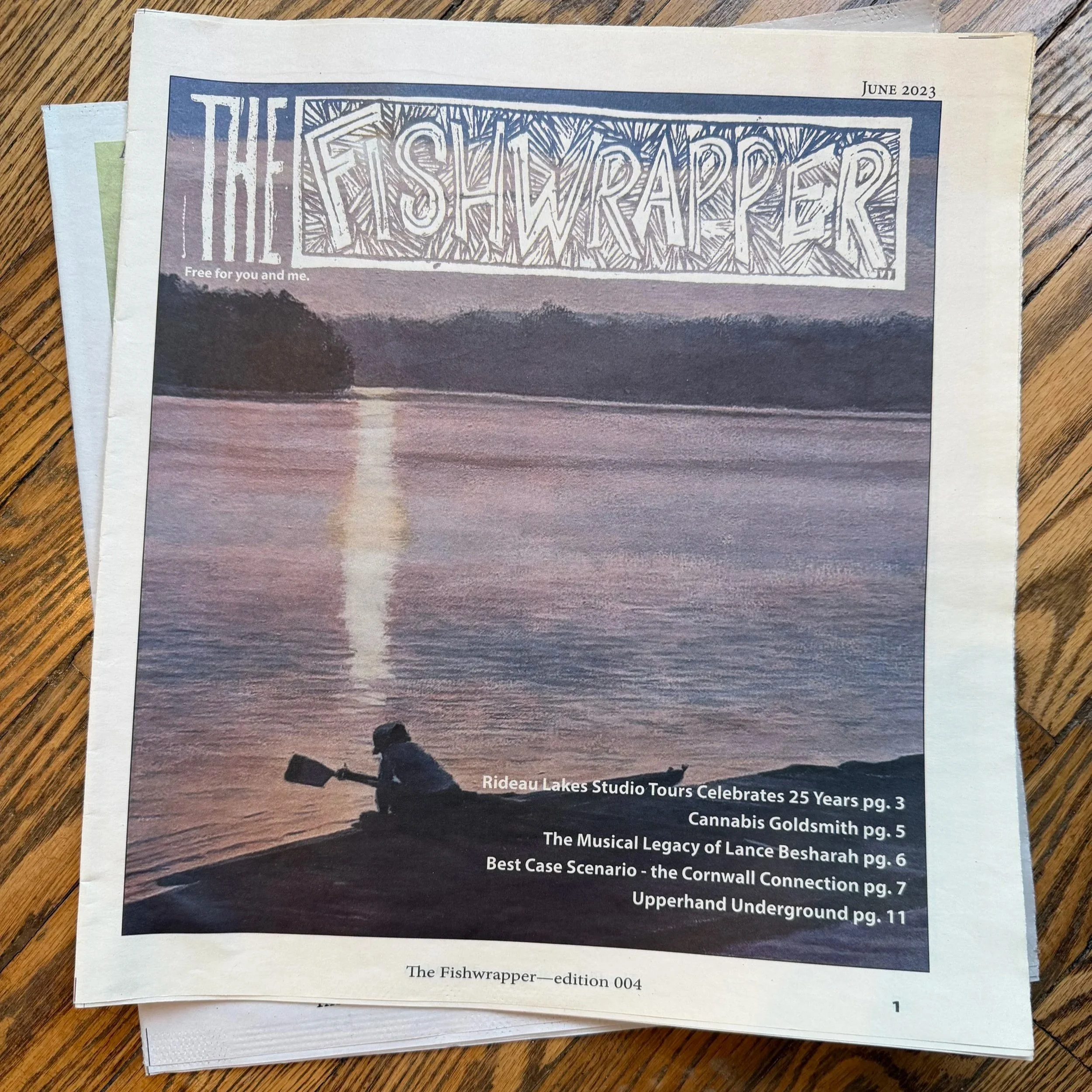 A newspaper titled 'The Fishwrapper' from June 2023 featuring a cover image of a person sitting in a boat on a calm lake at sunset, with the sky reflected in the water, and trees in the background. The cover includes headlines about local tours, cannabis, a musical legacy, and underground scenes.