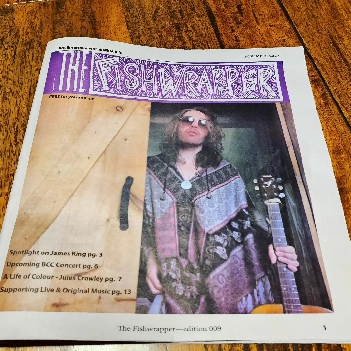 A newspaper titled "The Fishwrapper" with a purple header and a photo of a man with long hair, sunglasses, and a patterned shirt holding an acoustic guitar, on a wooden table.