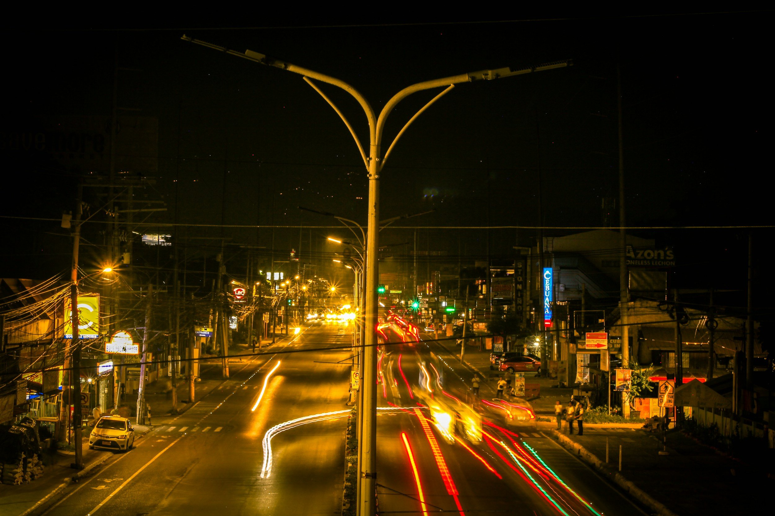 Nighttime city street with moving car lights and illuminated signs, and a large streetlamp in the foreground.