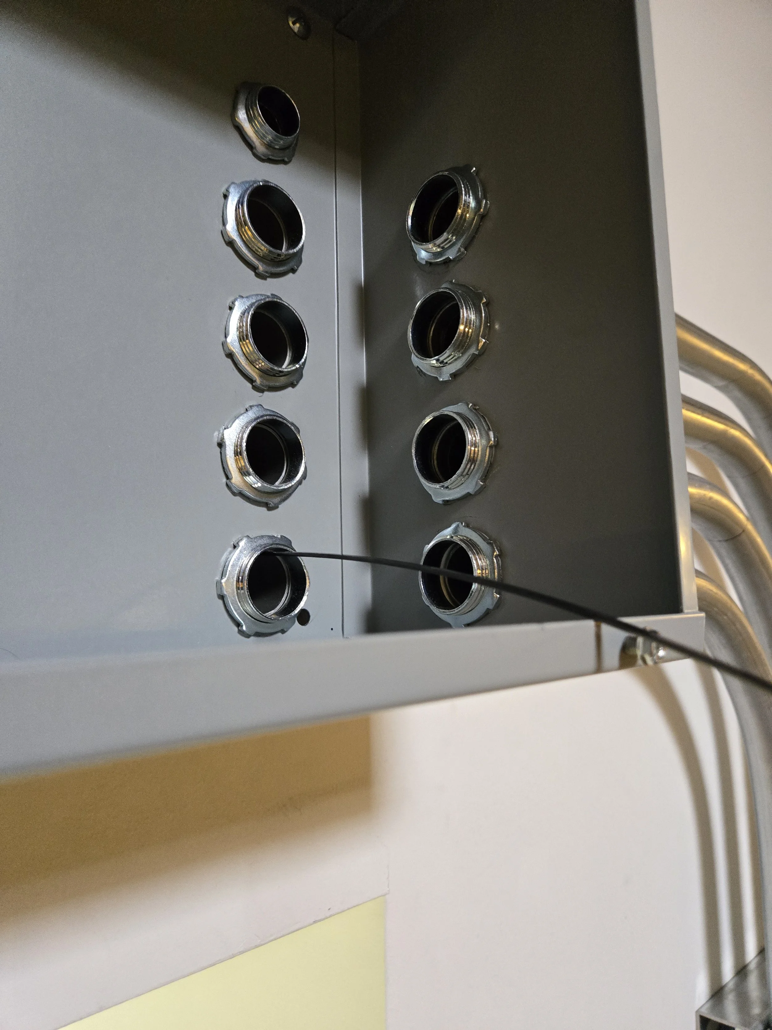Close-up of an electrical panel or junction box with multiple empty cable entry points, some with metal rings, and a metal wire protruding from one of the entry points. Several metal conduit pipes are connected on the right side.