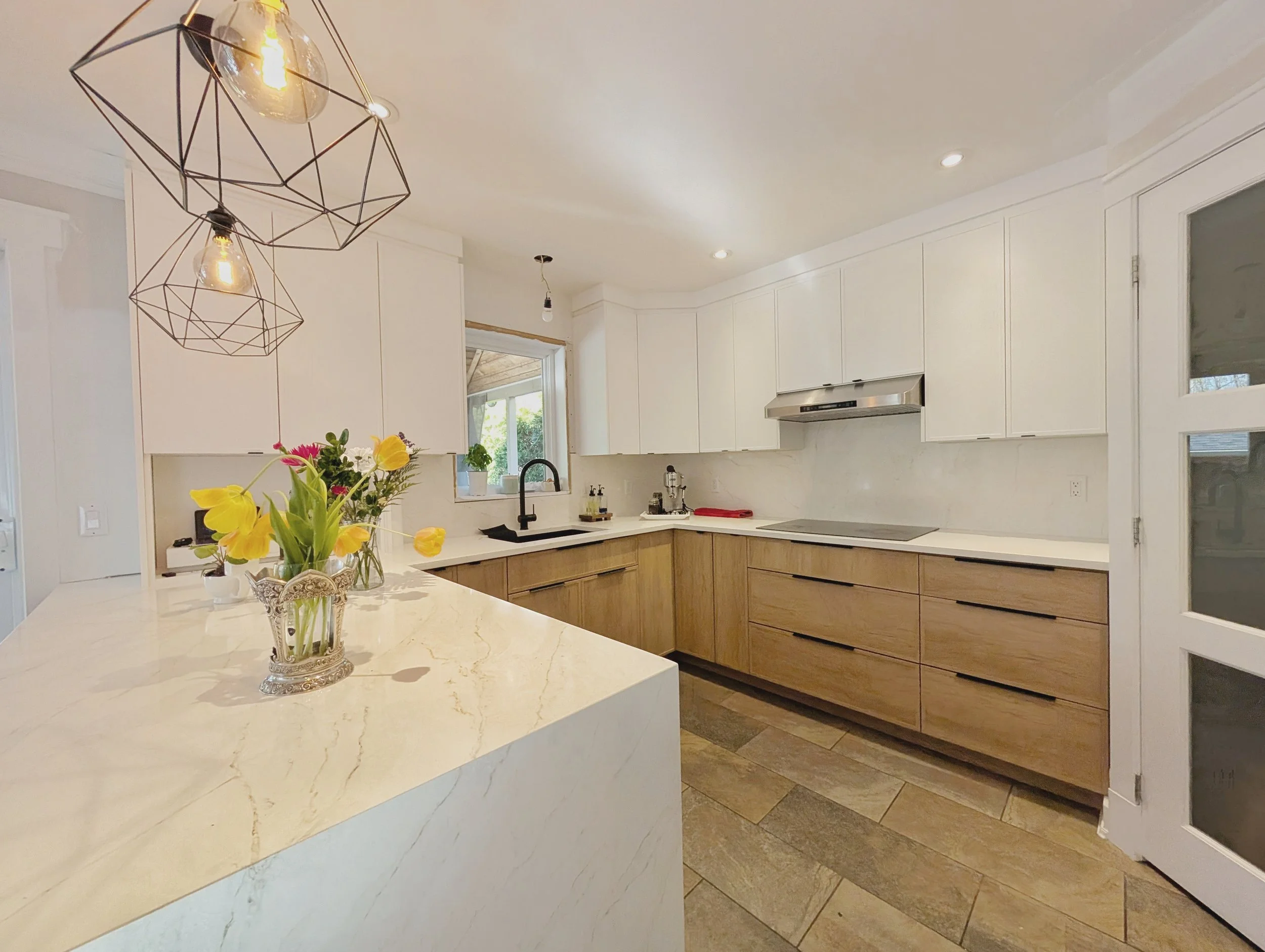 Modern kitchen with white upper cabinets, light wood lower cabinets, marble countertops, a black sink, and a stove. There are geometric pendant lights and a flower vase with yellow tulips on the island.