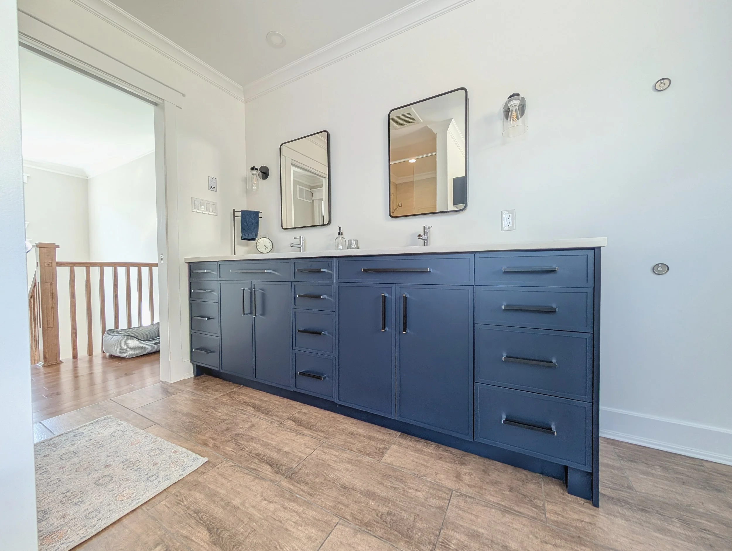 Bathroom with a navy blue vanity, two mirrors, and a double sink, in a bright room with white walls and wood flooring.