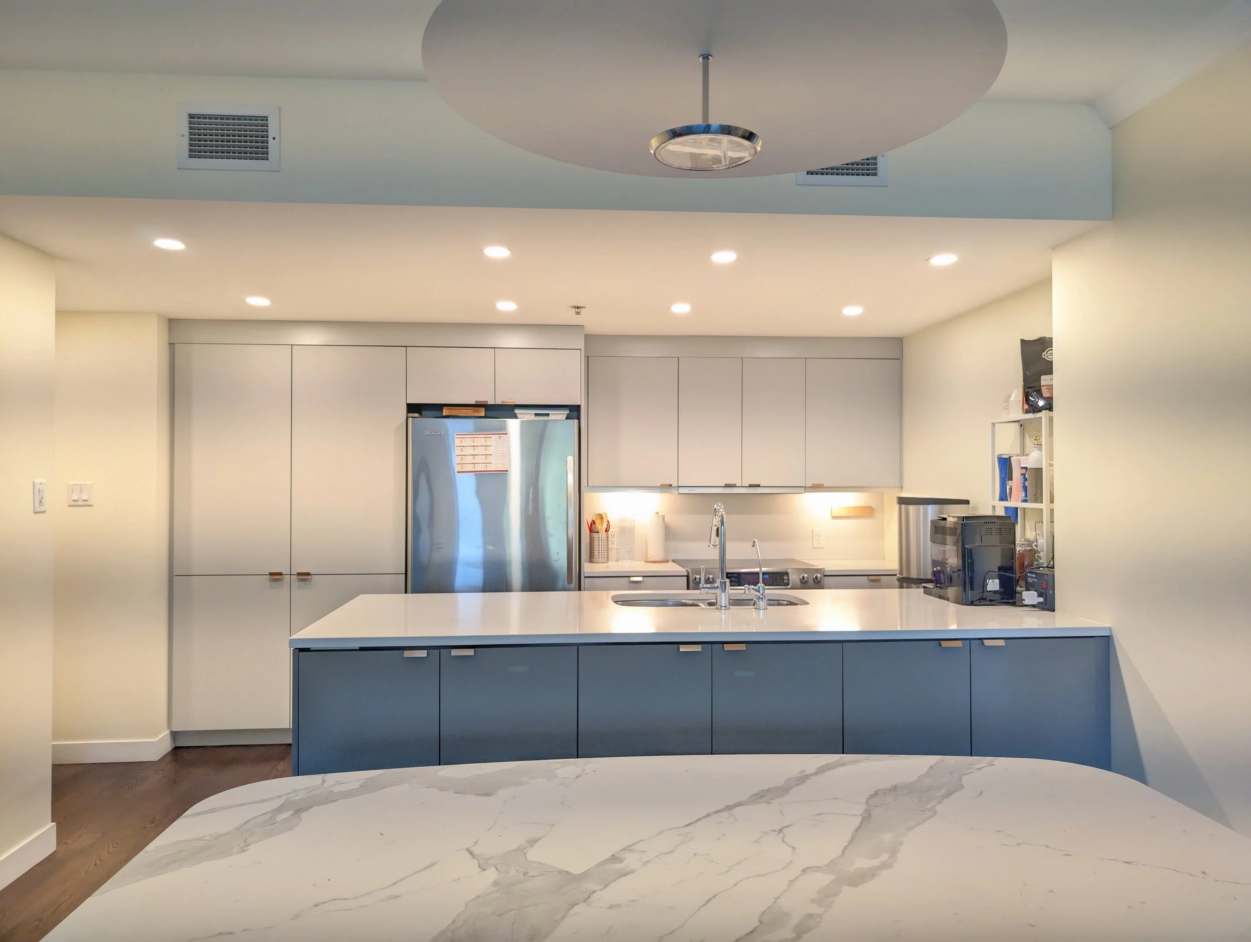 Modern kitchen with blue cabinetry, white countertops, stainless steel appliances, and a marble breakfast bar in the foreground.