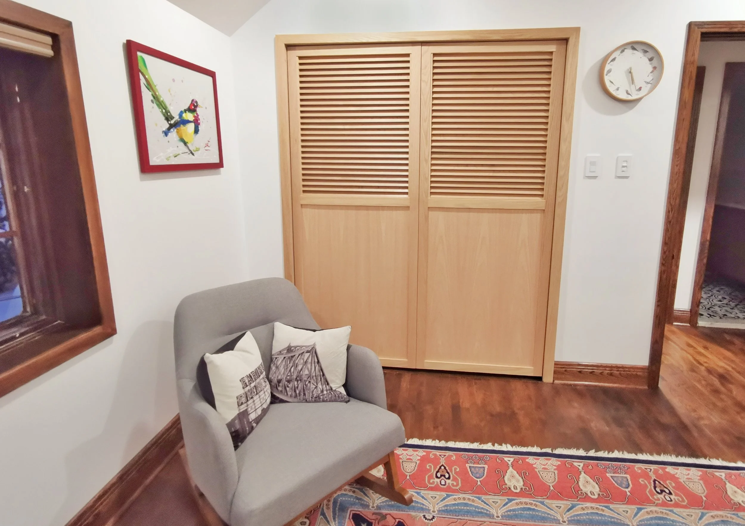 Living room corner with a light gray armchair, decorative pillows, a colorful abstract wall art, a wooden closet with louvered doors, a wall clock, a decorative rug, and hardwood floors.