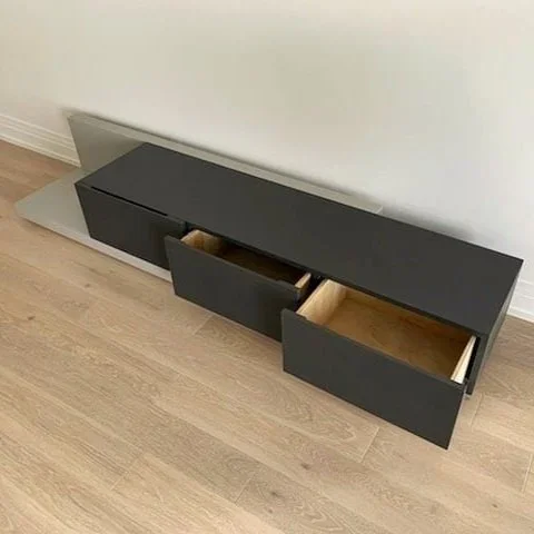 Black modern low-profile TV stand with open drawers on light wood floor, against a white wall.