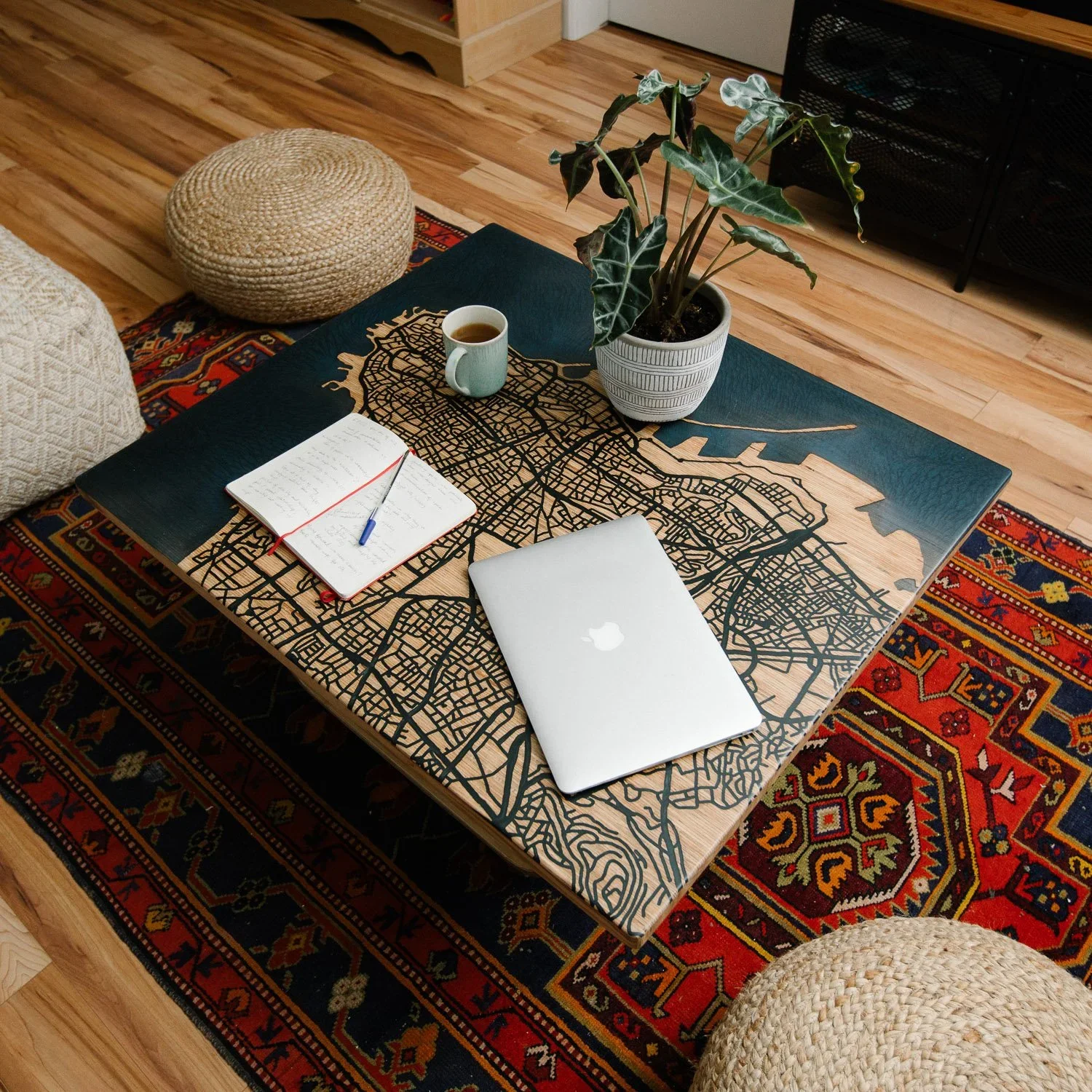 A wooden table with a city map design, a laptop, a notebook with a pen, a cup of coffee, and a potted plant. The table is on a colorful oriental rug, with woven poufs and wooden floors surrounding it.
