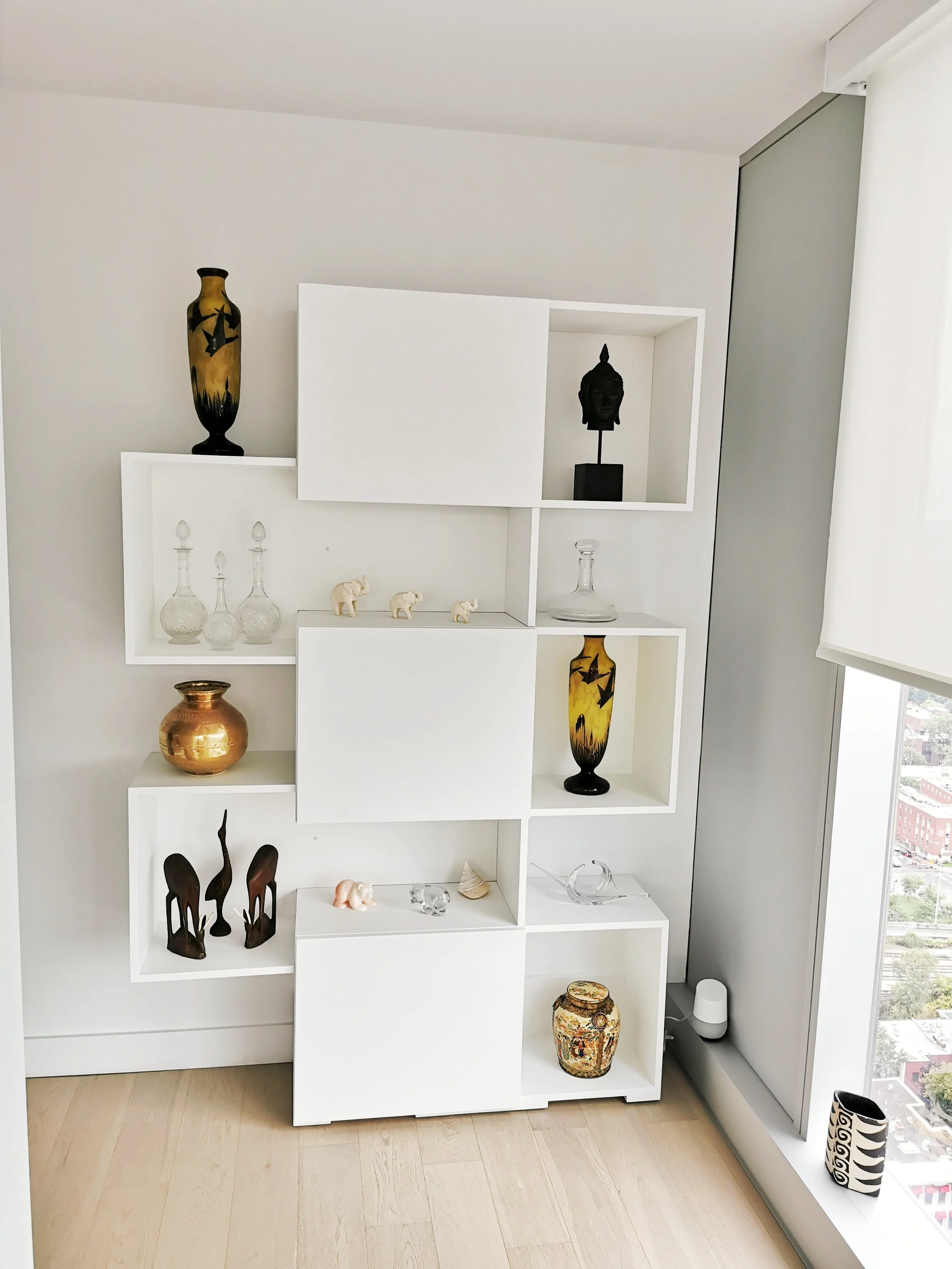 White modern shelving unit with decorative vases, statuettes, and sculptures, next to a large window with a city view