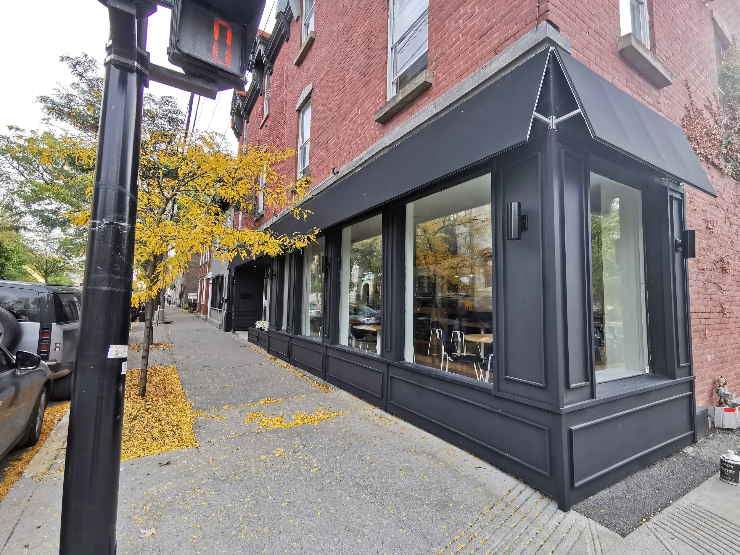 Corner of a brick building with black storefront and large glass windows, a small seating area inside, a canopy above the window, and trees with yellow leaves on a sidewalk with fallen yellow leaves, parked cars, and a street sign