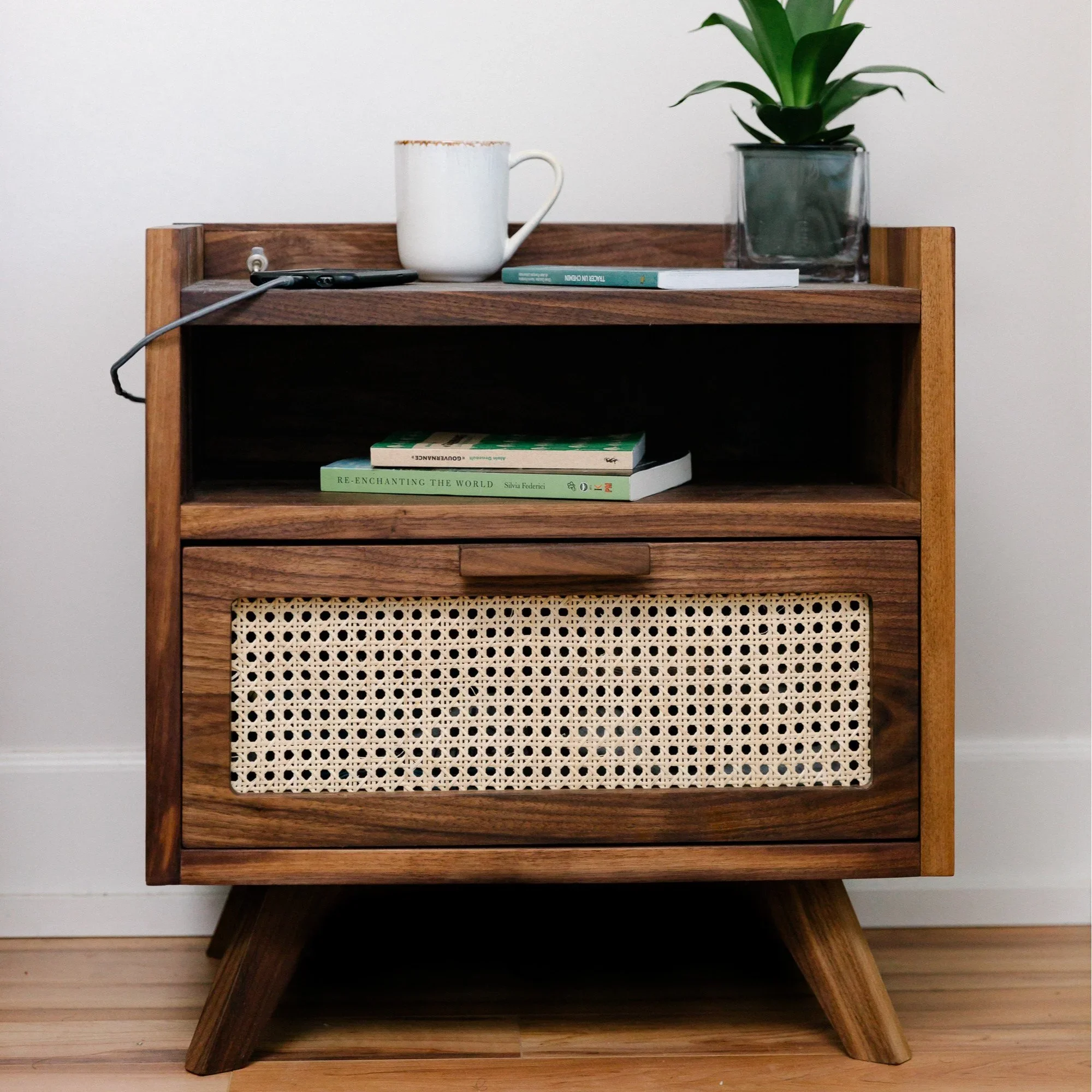 A mid-century modern wooden nightstand with a woven cane front drawer, three tapered legs, and an open shelf holding books. On top, there is a white mug, a potted plant in a transparent square pot, a book, and an earpiece with a wire.
