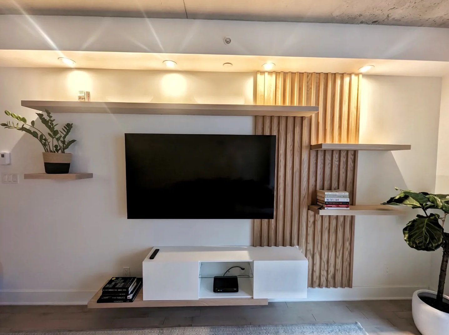 Modern living room with mounted flat-screen TV, wooden wall panels, white shelving, and potted plants.