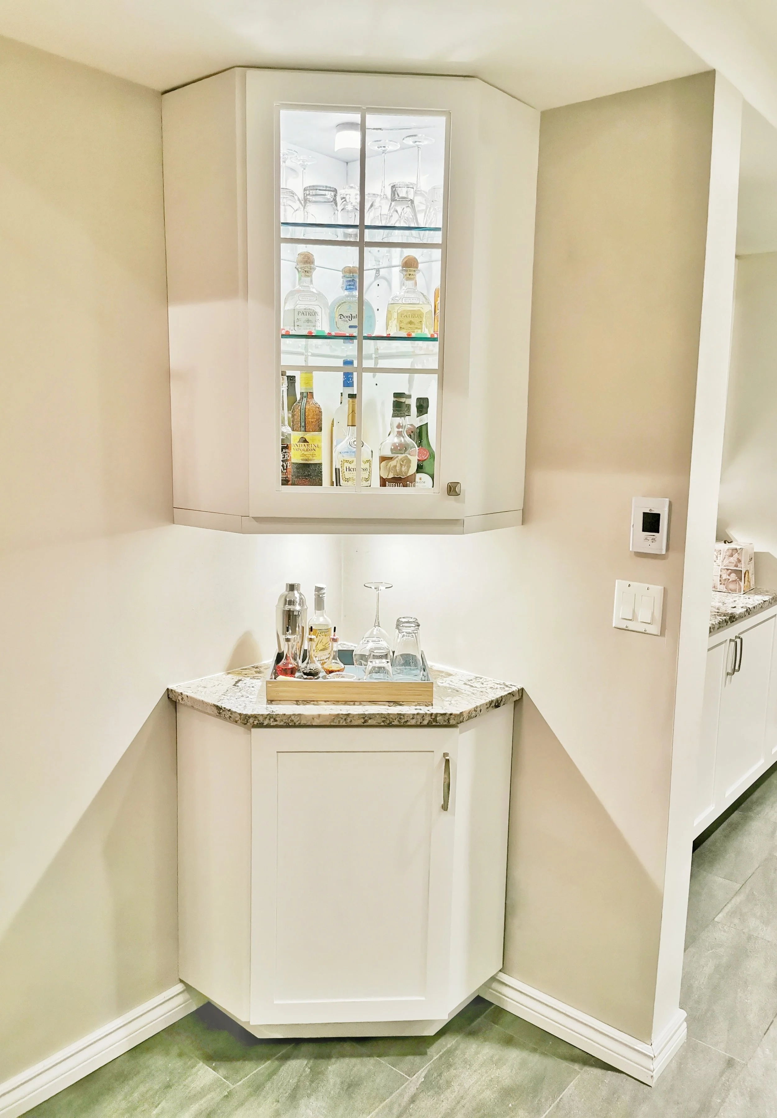 A corner bar area with a glass-fronted cabinet holding various bottles of alcohol and glassware, below a wall-mounted cabinet, all in a bright, modern kitchen.