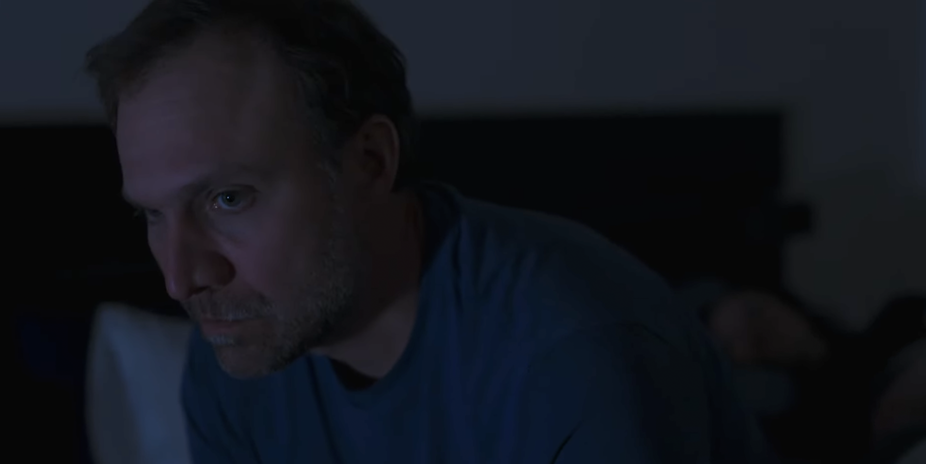 A middle-aged man with light-colored hair and beard, wearing a blue shirt, looking intently at something in a dimly lit room.