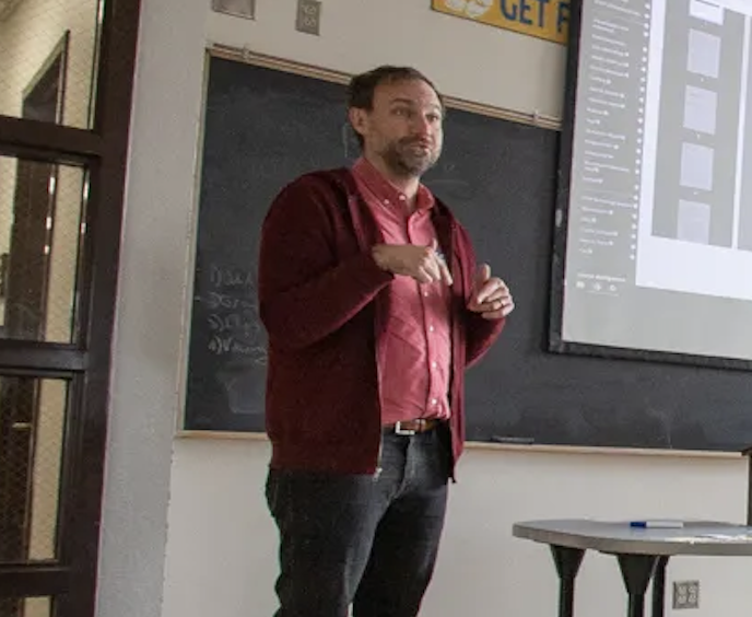 Man giving a presentation in front of a blackboard and a projector screen in a classroom.