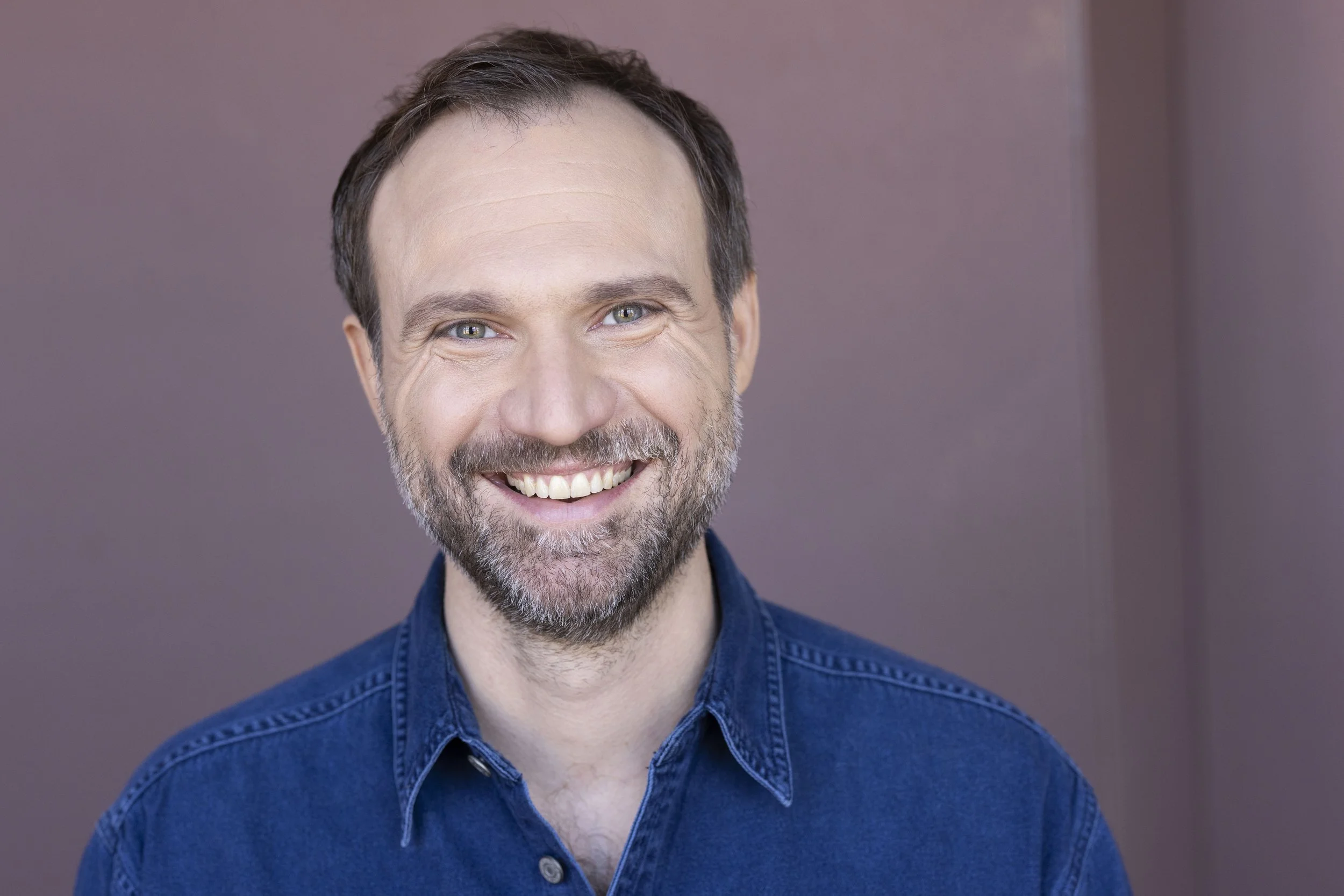 A smiling man with short brown hair, a beard, wearing a blue denim shirt, against a plain, muted background.