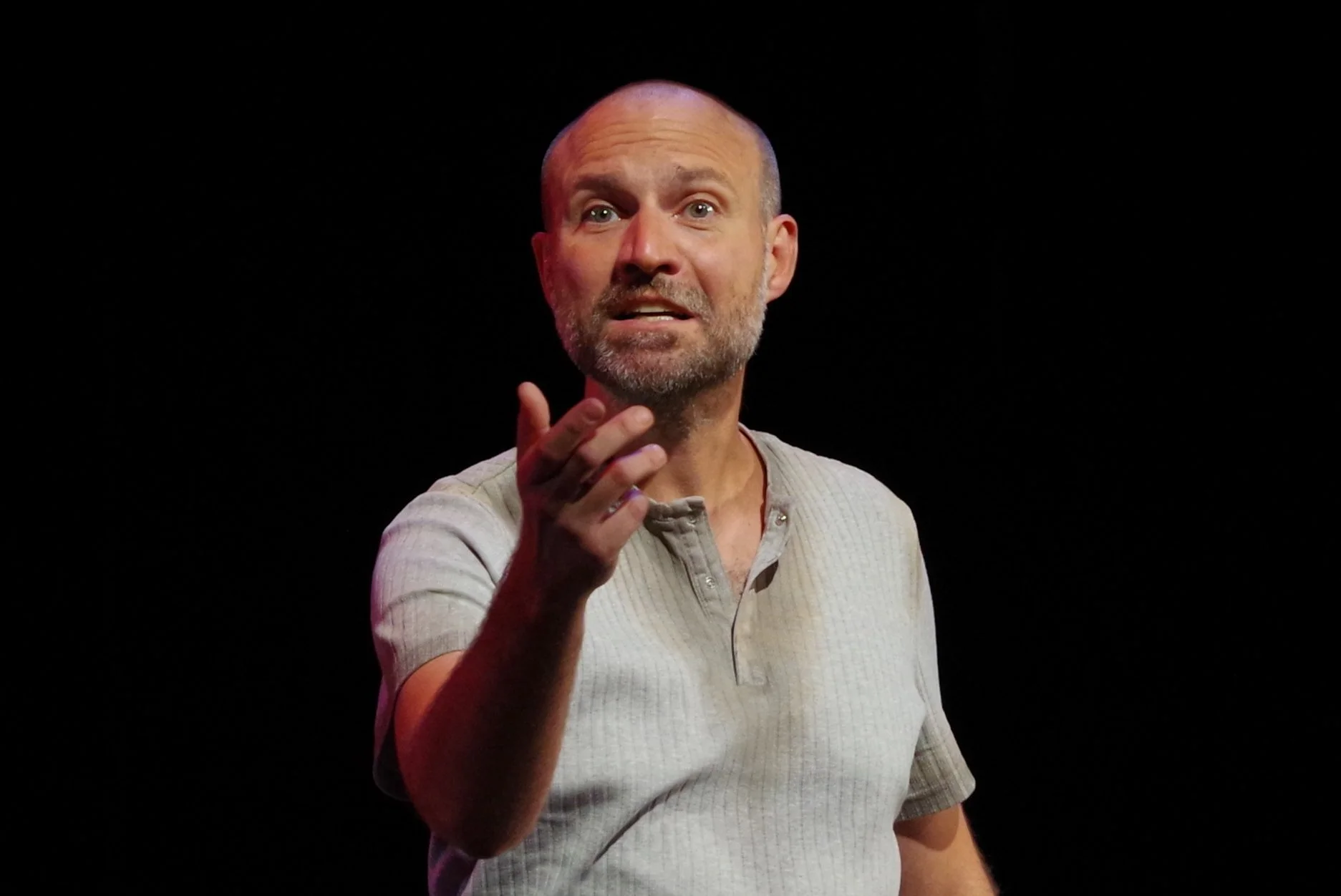 A man with a beard and short hair, wearing a light gray, short-sleeved, button-up shirt, is gesturing with his right hand while looking directly at the camera against a black background.