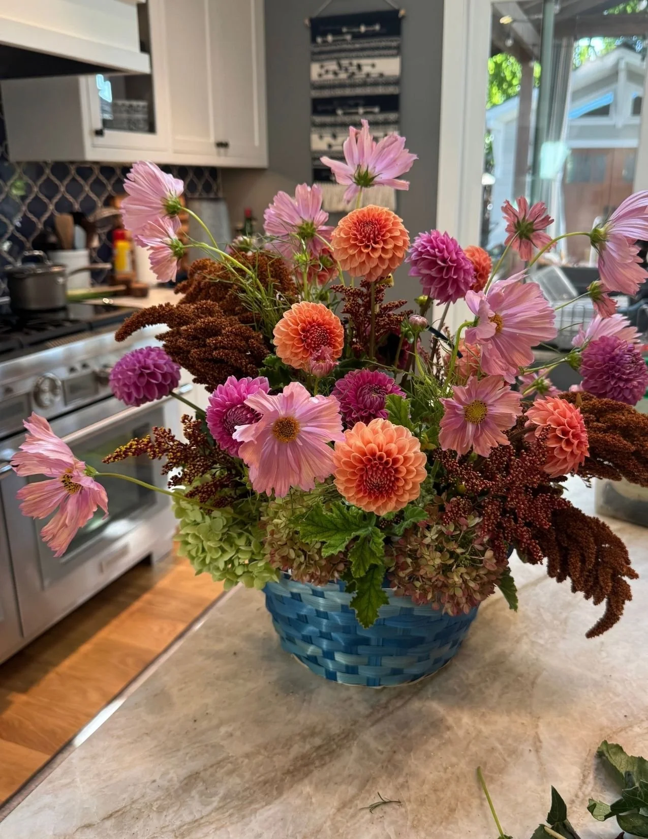 An abundant tabletop arrangement overflowing with dahlias, ranunculus, and deep wine-colored floral accents, beautifully arranged in a woven blue basket with a handle.