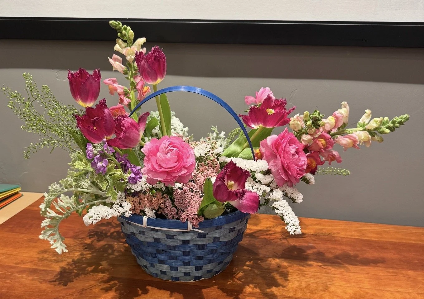 Fresh garden-style floral basket woven in dark blue wicker, filled with hot pink roses, purple stock, and airy white filler flowers.