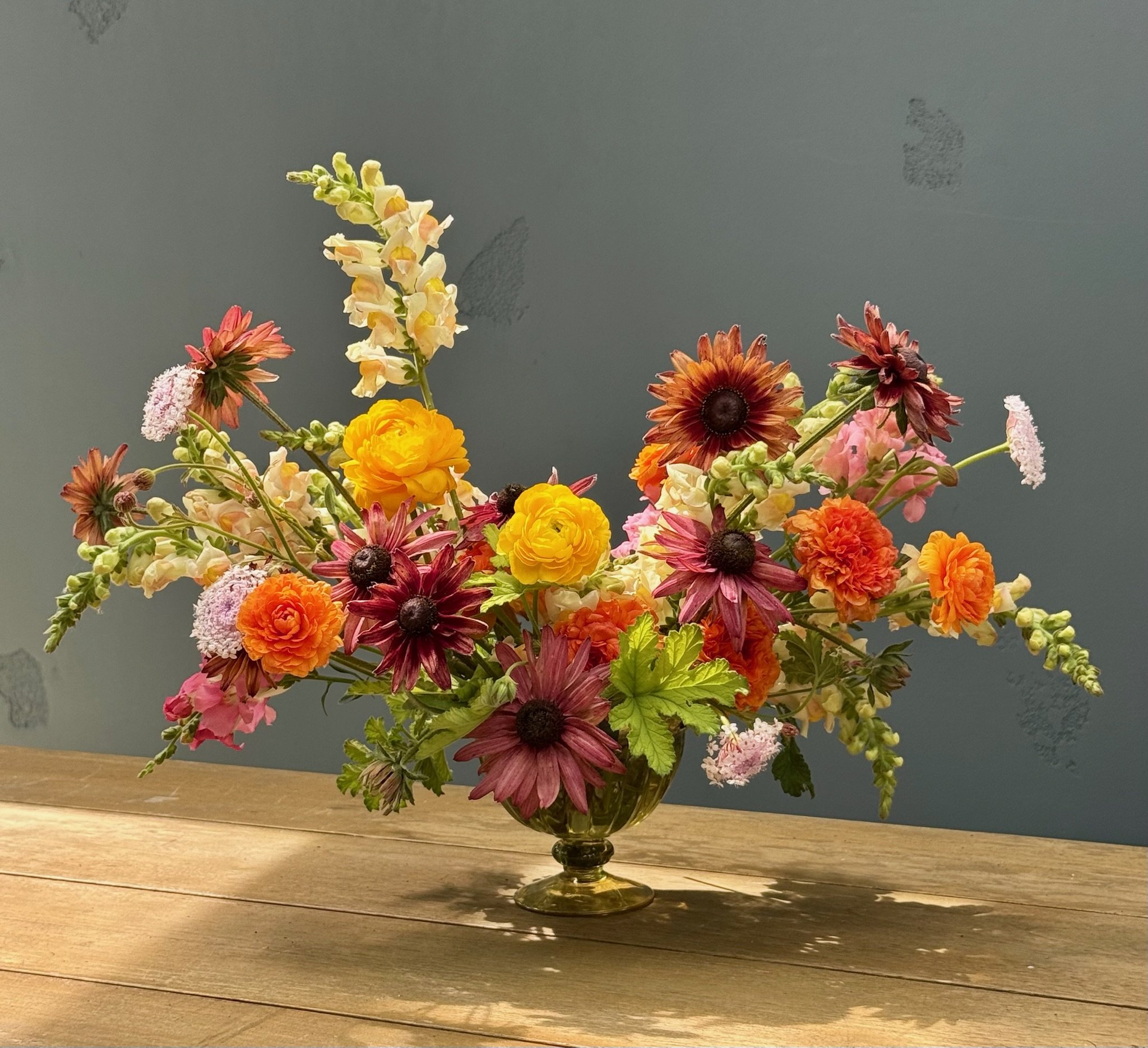 Vibrant floral arrangement with orange, pink, and yellow flowers in a golden footed pedestal bowl.