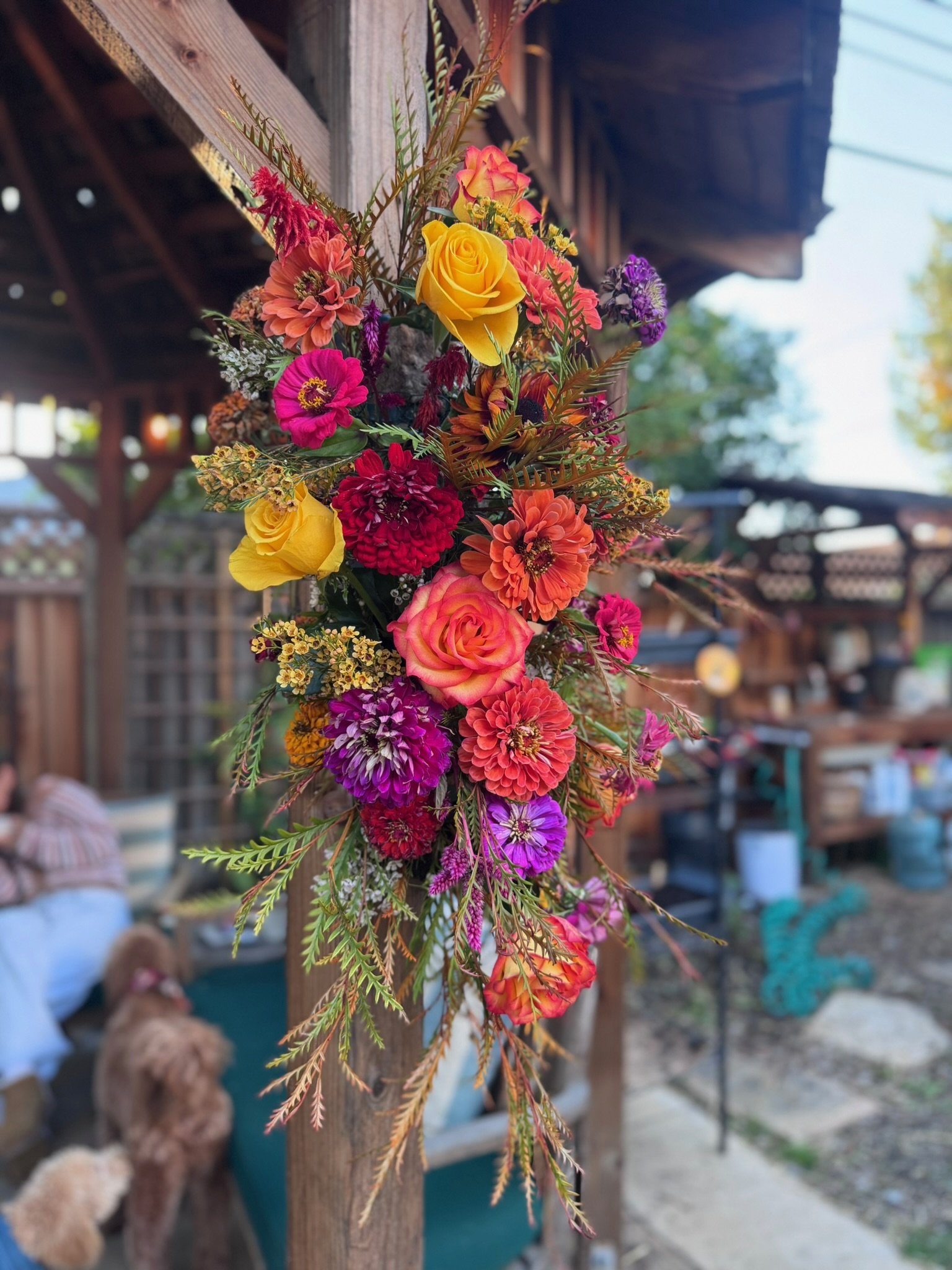 Colorful artificial flower arrangement hanging outdoors with pink, yellow, orange, purple, and red flowers among green foliage, attached to a wooden structure in a backyard.