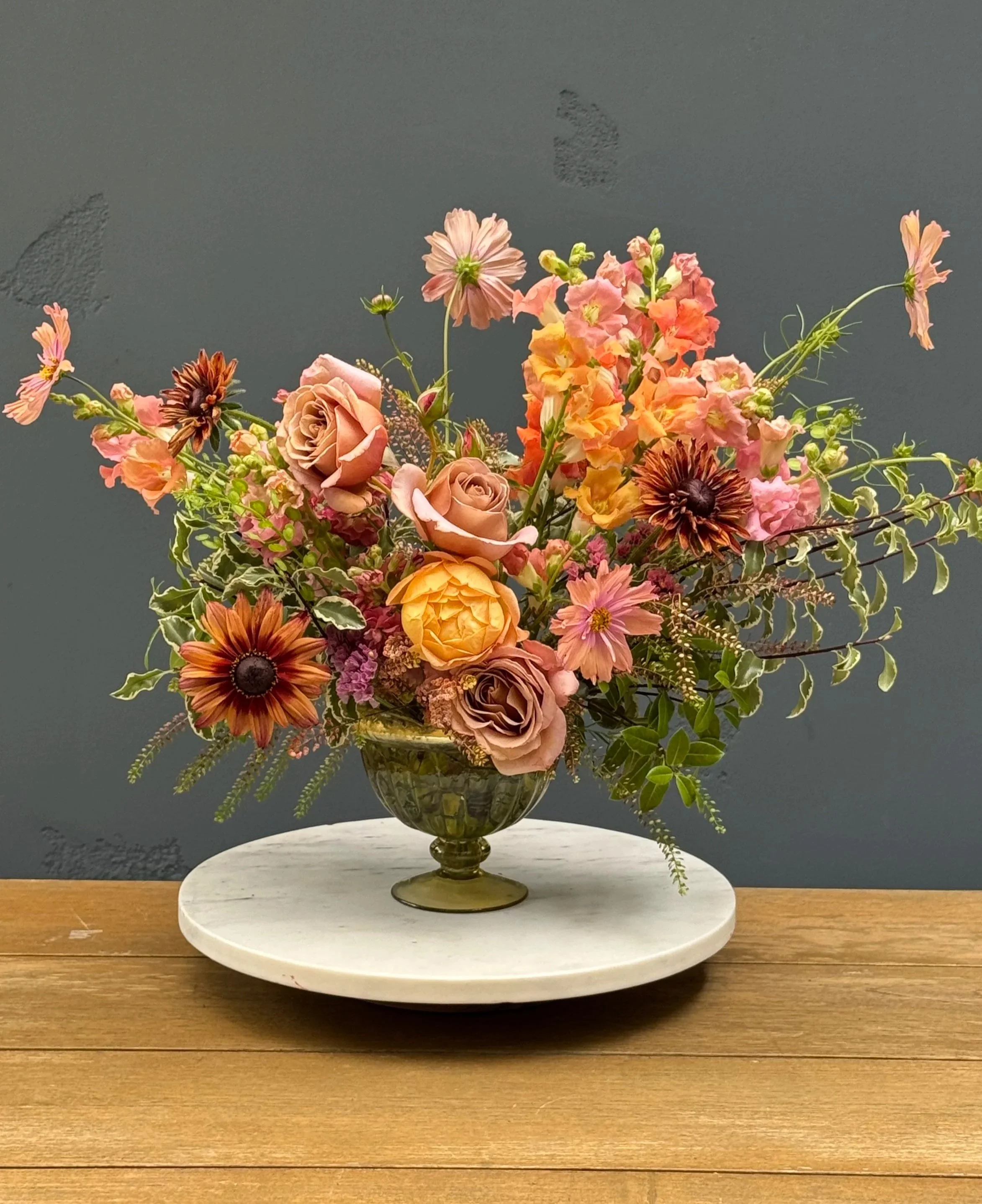 A floral arrangement in a glass vase on a white marble pedestal, featuring roses, dahlias, and other flowers with green foliage, on a wooden surface against a dark background.