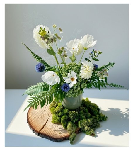 White and blue floral arrangement with greenery, displayed in a mint green vase set on a natural wood slice backdrop.