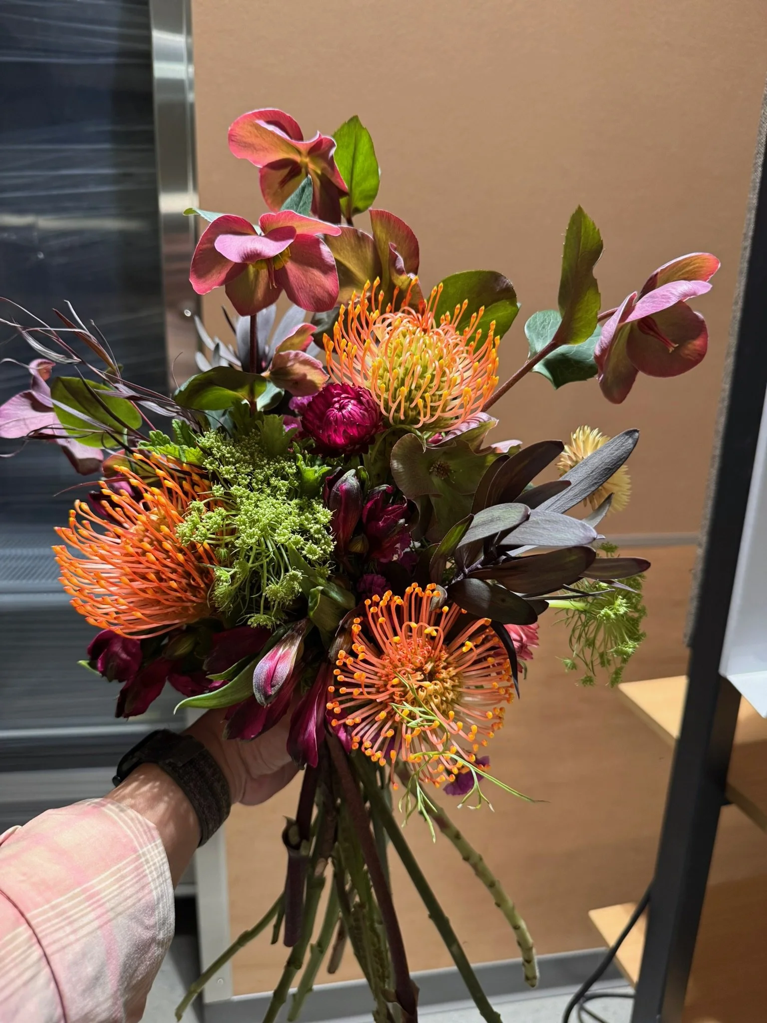 A hand holding a colorful bouquet of flowers, including pink and orange blooms with green foliage, against a background of a beige wall and part of an elevator.