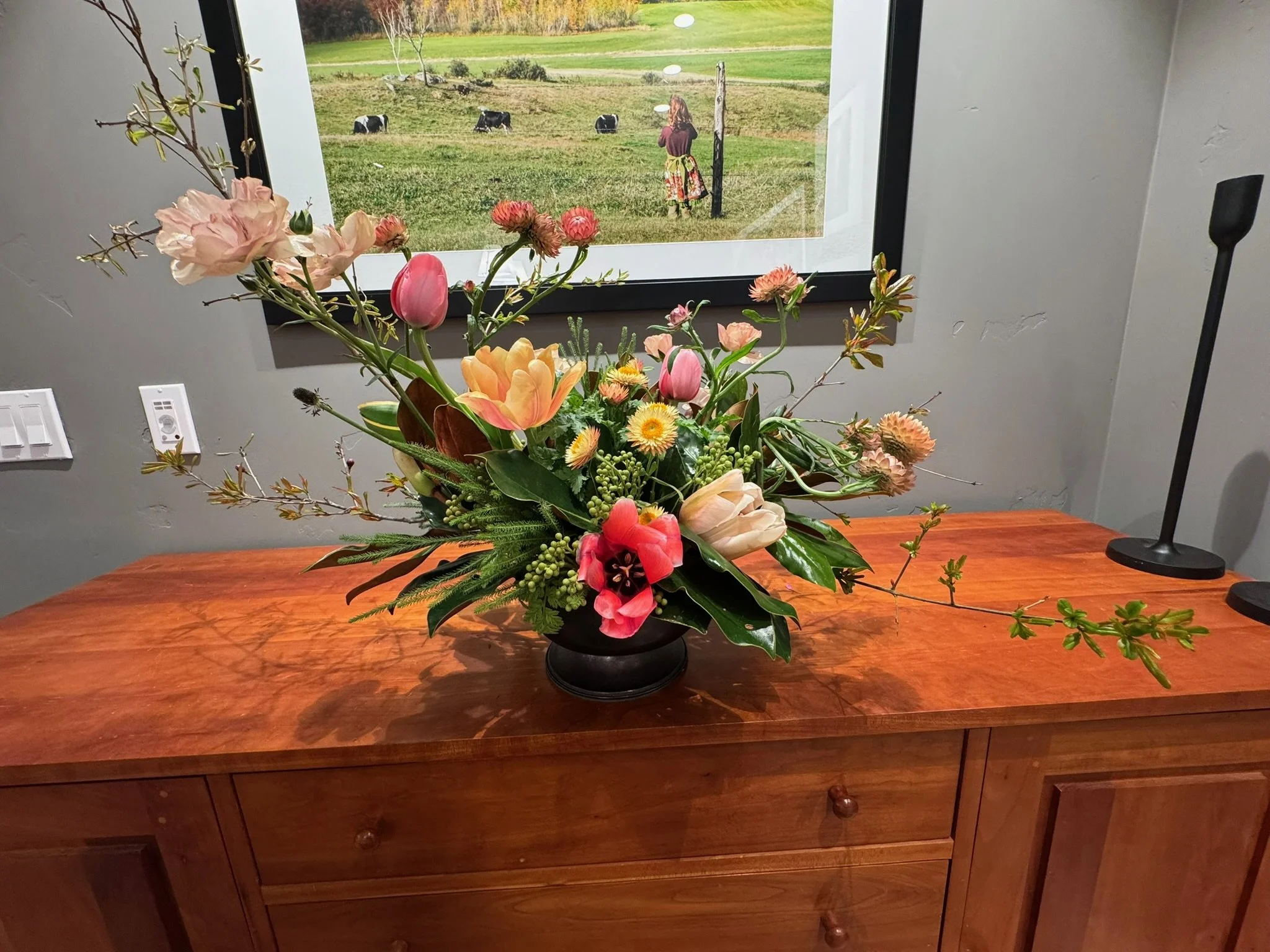 A wooden table with a large colorful flower arrangement in a black vase, set in front of a window showing a woman standing outdoors in a grassy field with cows grazing.