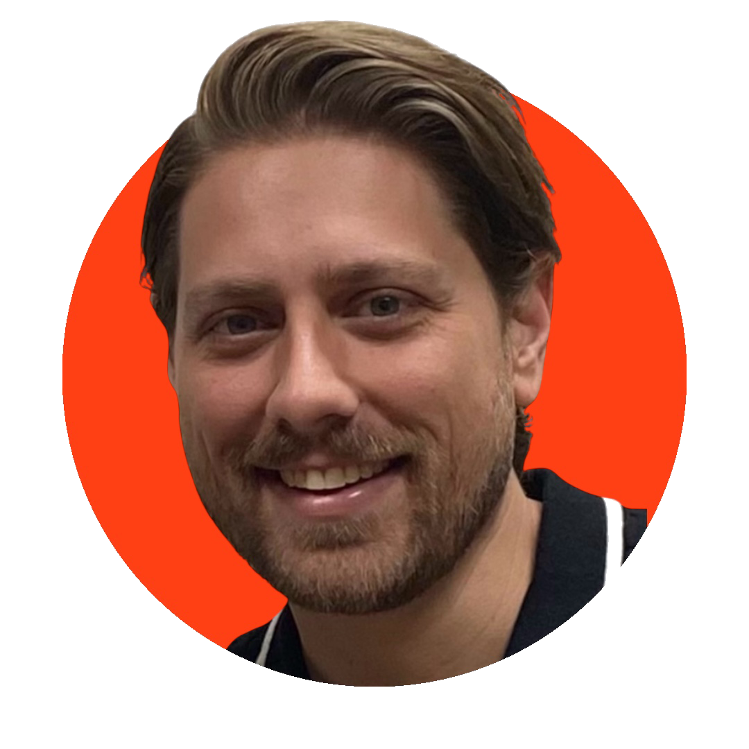 A smiling man with light brown hair, a beard, and a mustache, wearing a dark shirt, against a red circular background.