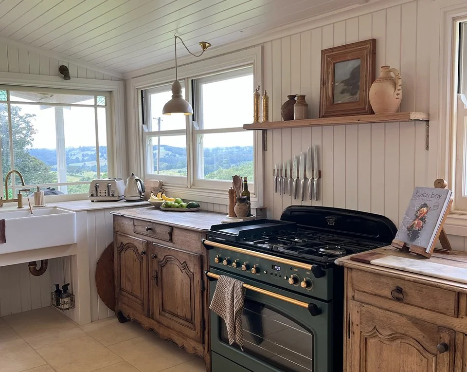 Rustic kitchen with white shiplap walls, a farmhouse sink, window with a farm view, wooden cabinets, a black stove with a towel hanging, a countertop with a toaster, kettle, and fruit, and decorative jars and artwork on shelves.