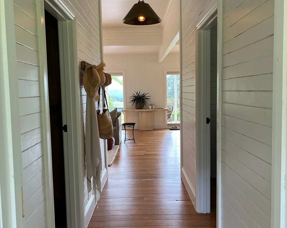 A bright hallway leading to a sunlit room with large windows, a desk, a plant, and wooden flooring.