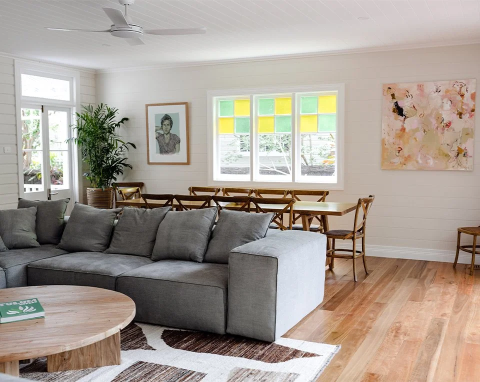 Bright living room with a sectional gray sofa, a round wooden coffee table, a potted plant, artwork on walls, and a dining area with wooden chairs and a table near colorful stained glass windows.