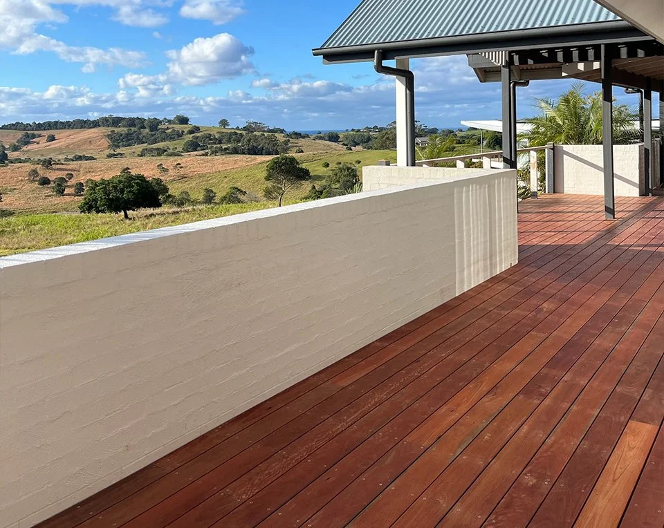 A spacious outdoor balcony with a wooden deck and a white brick wall, overlooking a scenic landscape of rolling hills with trees and grass, under a partly cloudy sky.