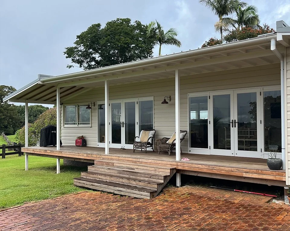 A house with a wooden deck, two sliding glass doors, and patio furniture, surrounded by green grass and trees.