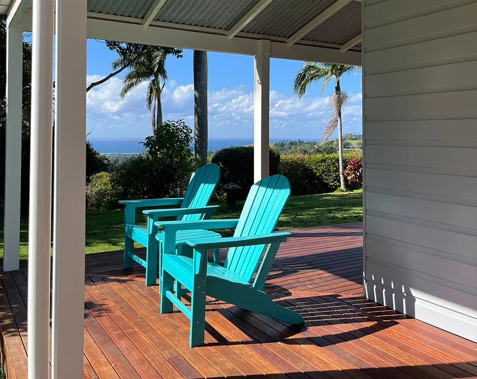 Two turquoise Adirondack chairs on a wooden porch overlooking a lush green landscape with palm trees and a view of the ocean under a partly cloudy sky.