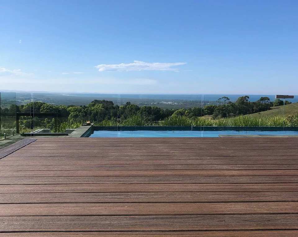 View of a swimming pool with a glass fence, overlooking a green landscape and the horizon under a blue sky.