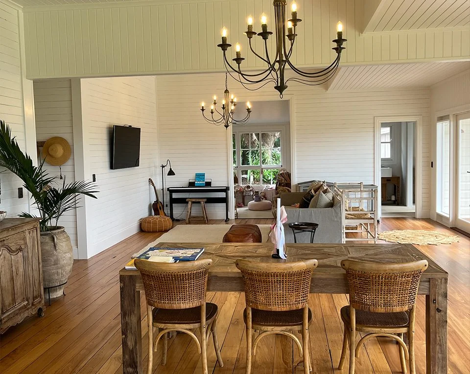 Open-concept living room and dining area with white paneled walls, wooden flooring, a large wooden dining table with four rattan chairs, a chandelier, a sofa with pillows, a small desk with a chair, a guitar, and large windows with views of greenery outside.