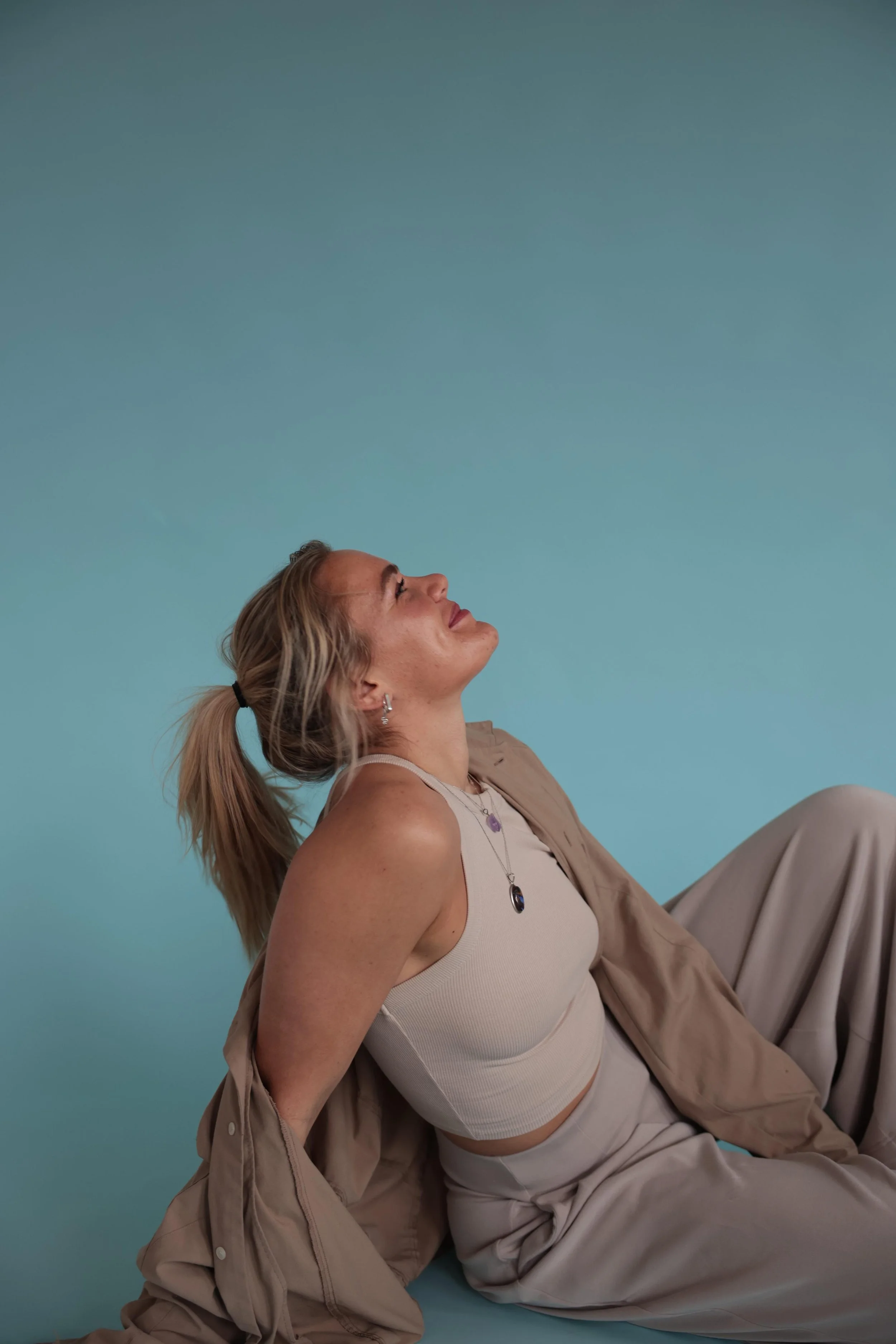 Een vrouw met blond haar in een paardenstaart, zittend op de grond tegen een lichtblauwe achtergrond, gekleed in beige kleding en sieraden, met haar hoofd naar achteren gekanteld en haar ogen dicht.