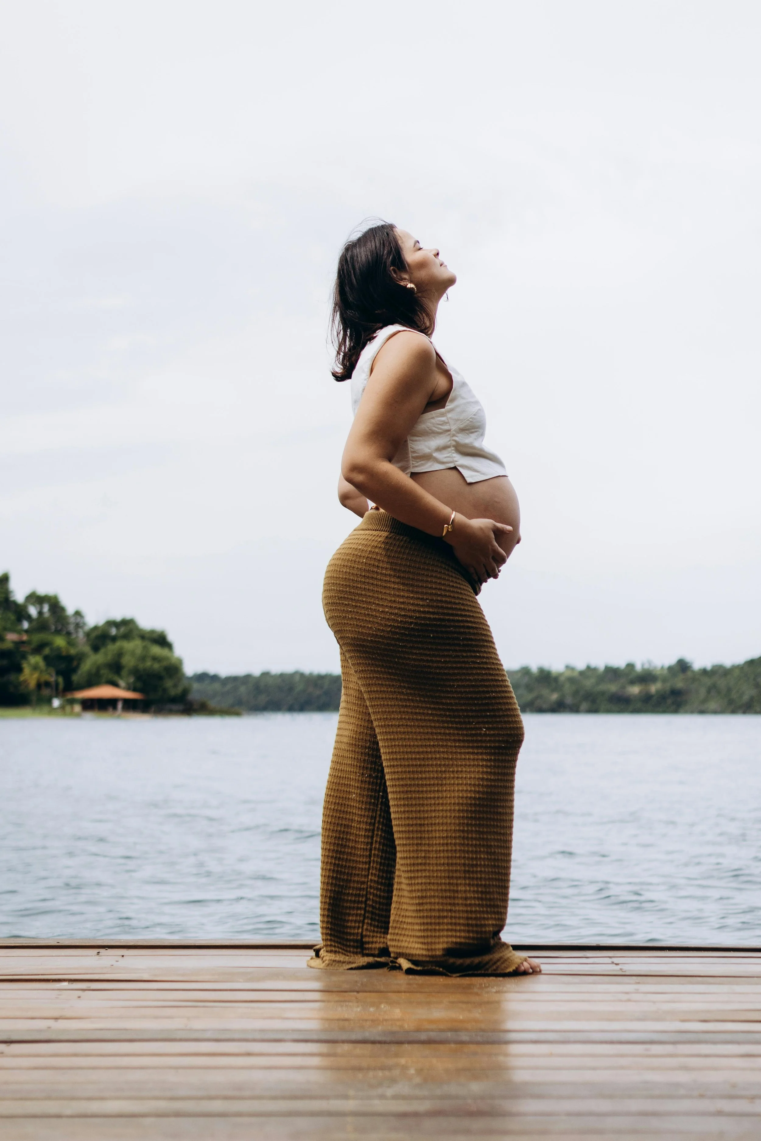 Zwangere vrouw staat op een houten dek aan de waterkant, kijkt omhoog, met water en groene bomen op de achtergrond.