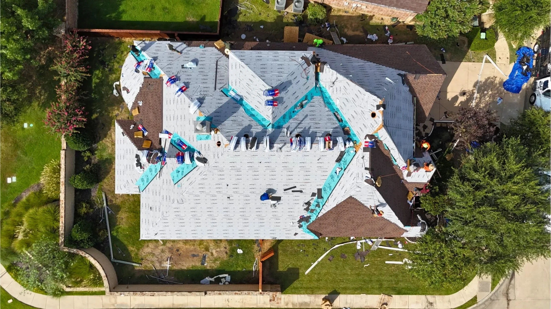 Aerial view of a house roof under construction with workers installing new roofing materials. The roof is partially covered with white underlayment, blue tarps, and roofing shingles, and surrounding the house are trees, a sidewalk, and parked cars.