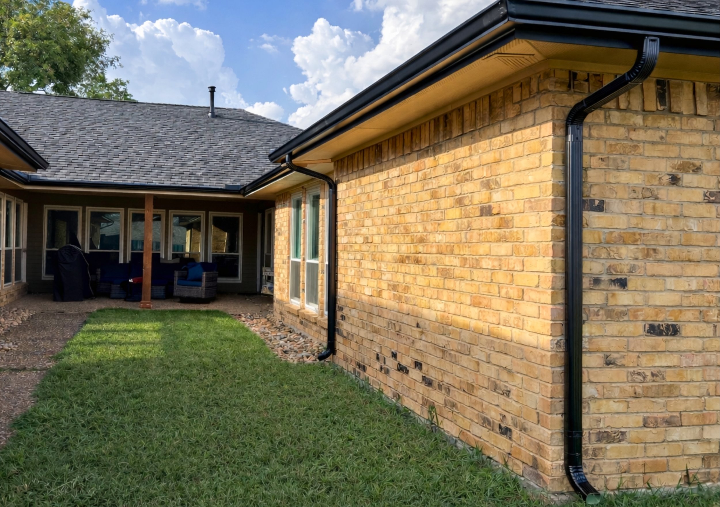 Backyard patio area with outdoor seating, brick house wall, black gutters, and green lawn under a blue sky with clouds.