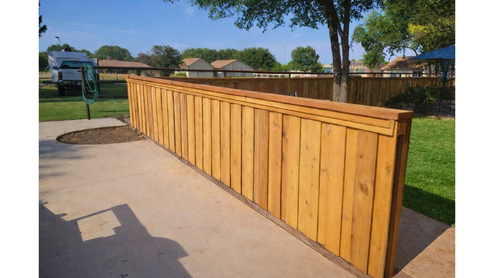 Newly built wooden privacy fence installed along a concrete patio in a backyard with trees, grass, and outdoor furniture.