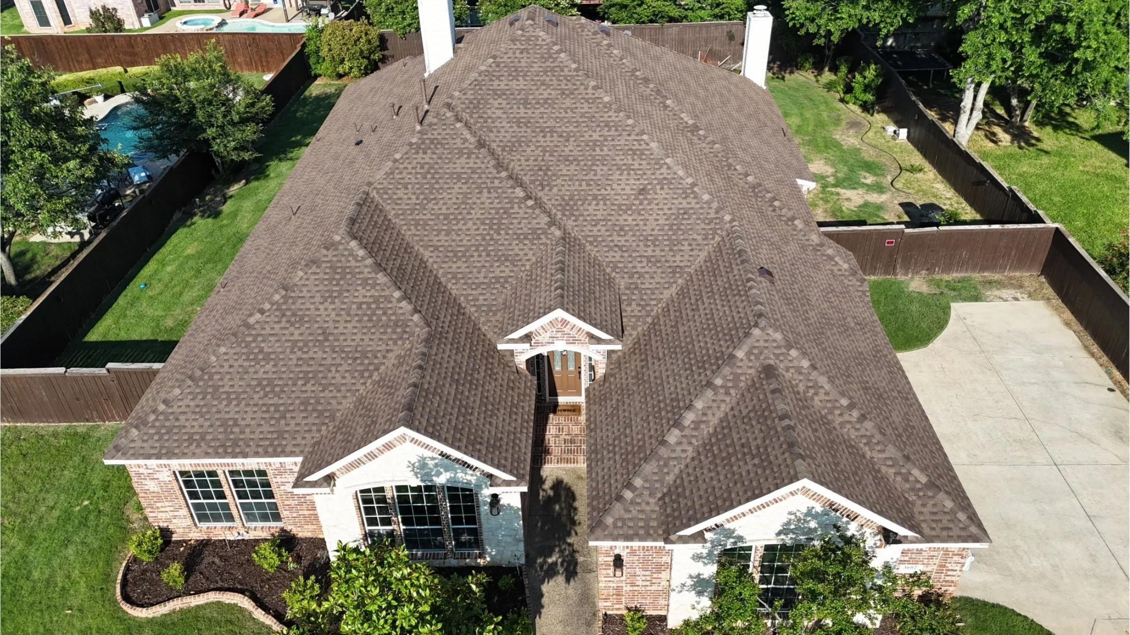 Aerial view of a suburban house with a brown shingle roof, brick walls, large windows, a front yard with landscaped plants, and a concrete driveway leading to a garage, surrounded by a fenced yard with trees and neighboring houses.