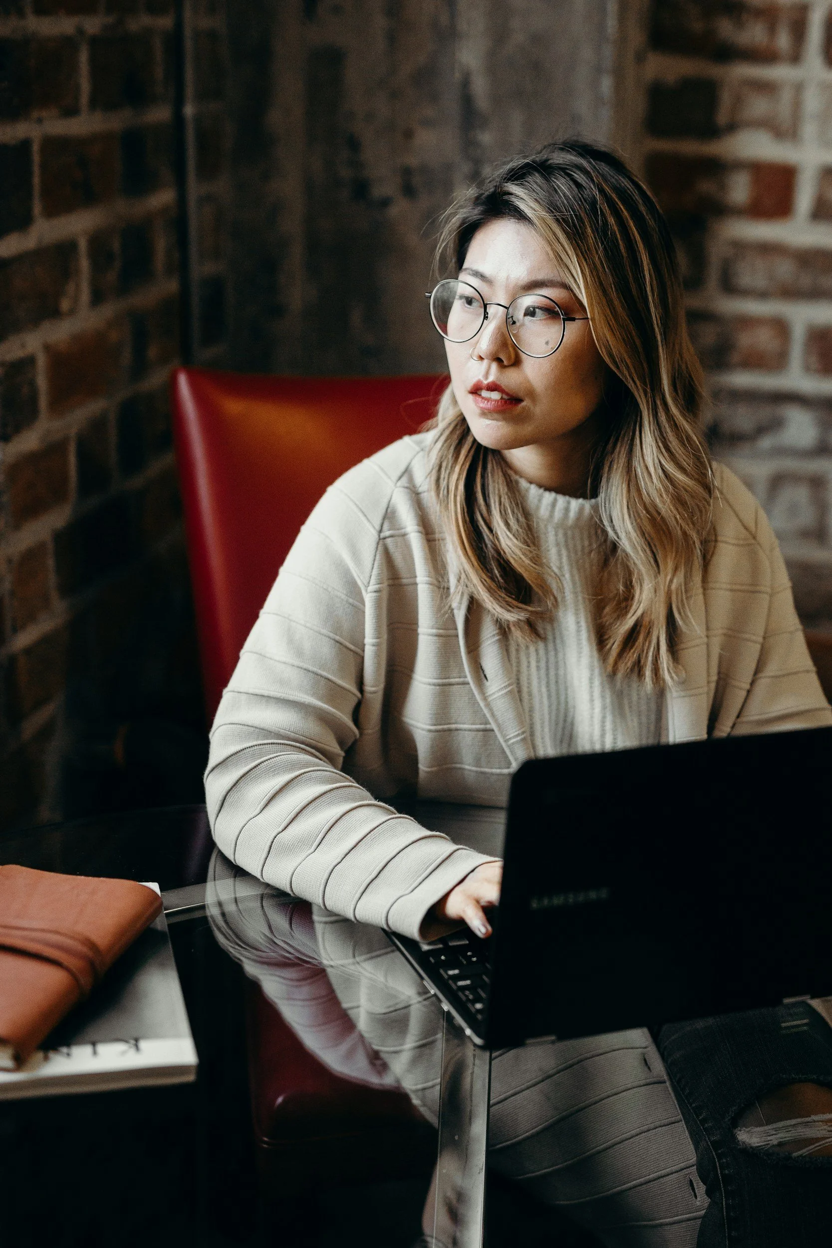 A woman with glasses and wavy hair working on a laptop at a table in a cozy, brick-walled cafe.