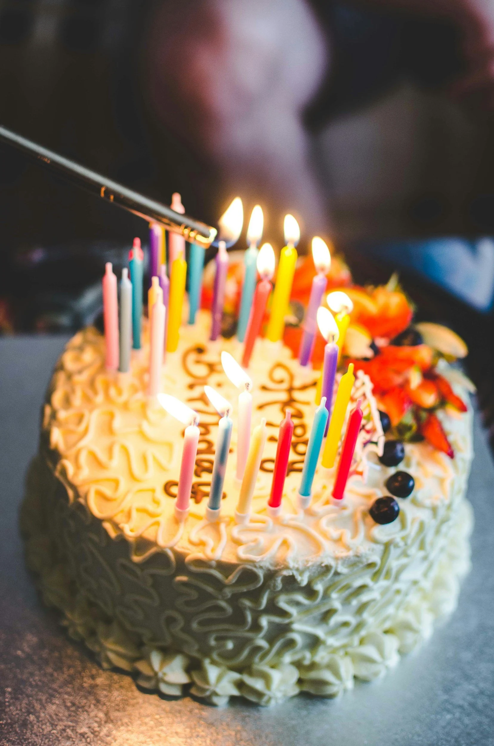 A decorated birthday cake with lit candles and a hand using a blowtorch to light them.