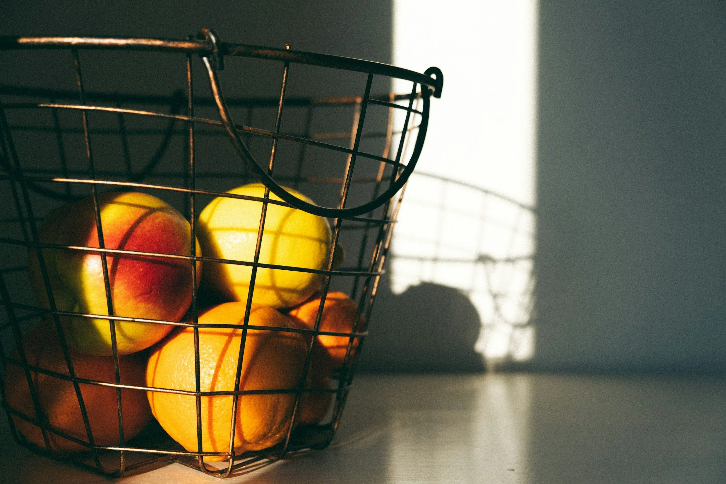 A wire basket filled with apples and oranges, with sunlight casting a shadow of the basket and fruits on a wall.