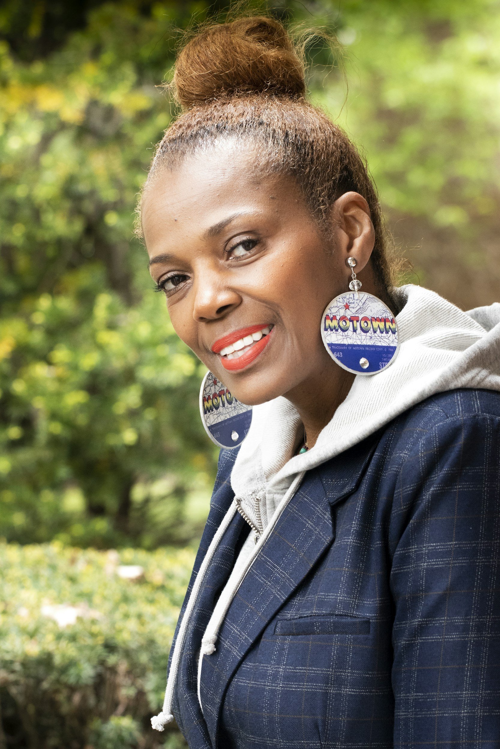 A woman with braided hair pulled into a bun, wearing large circular earrings with 'Motown' written on them, a hoodie, and a plaid blazer, smiling outdoors with blurred green foliage in the background.
