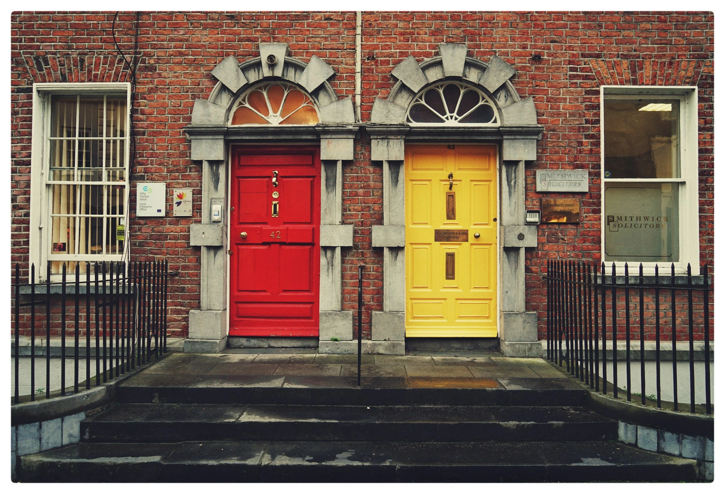 Two brick townhouse doors, one red and one yellow, with arched windows above, black iron railings, and steps leading up to each entrance.