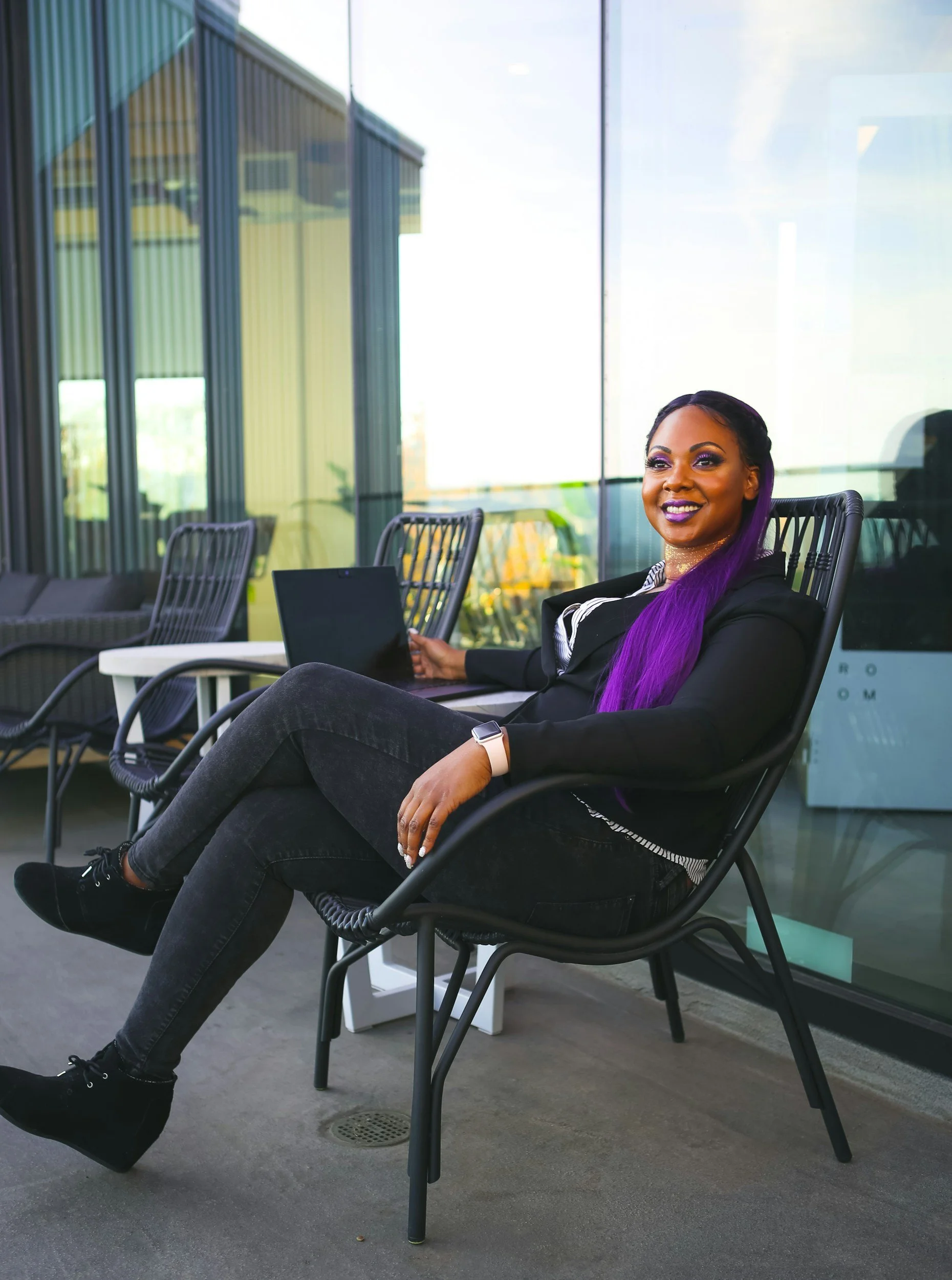 A woman with purple hair sitting comfortably with a laptop outdoors on a patio, smiling, in front of glass windows reflecting the sky and surrounding buildings.