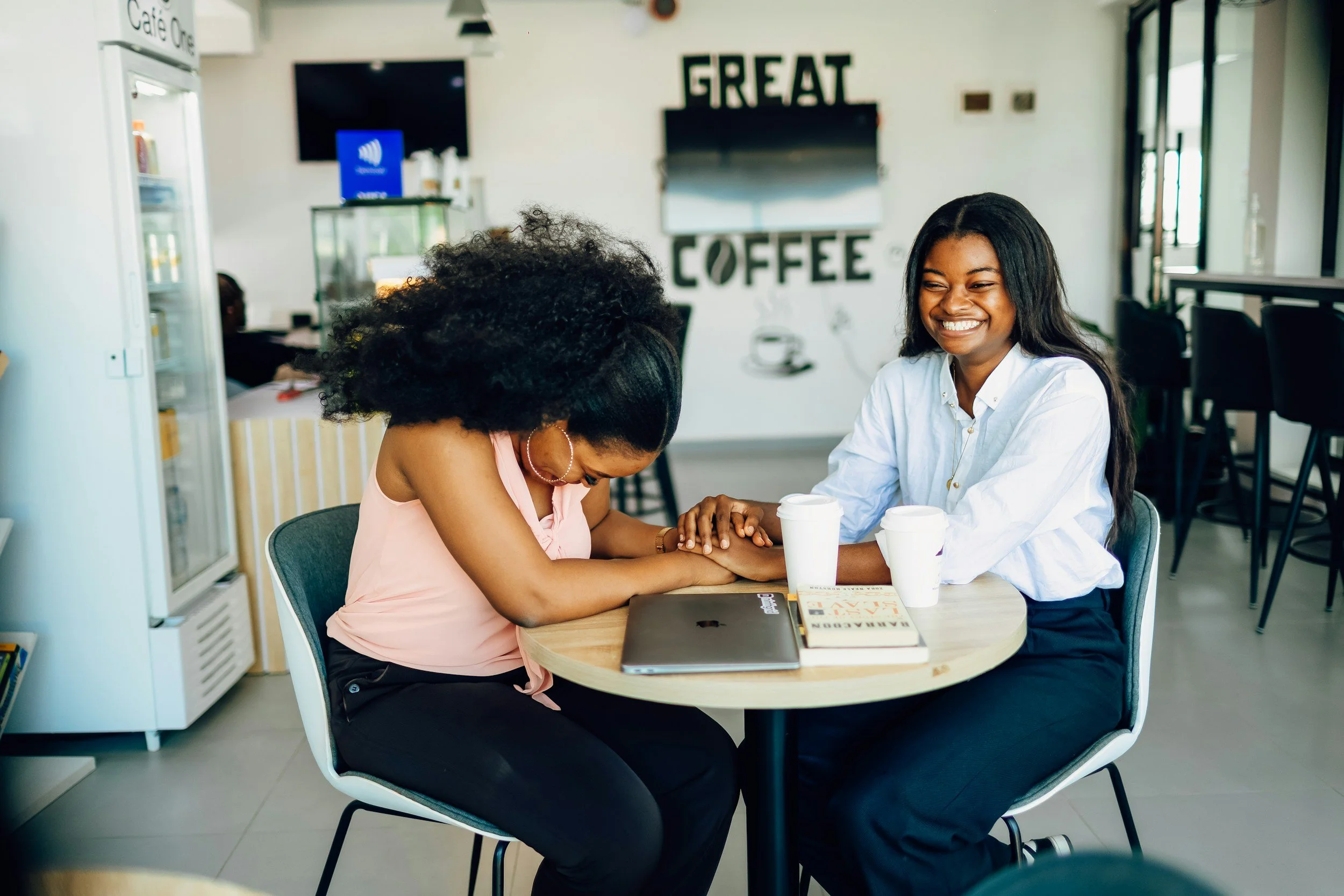 Two women sitting at a table in a coffee shop, engaging in a heartfelt moment with one woman holding the other's hand and both smiling; coffee cups and a laptop on the table, with a sign on the wall behind that reads 'GREAT COFFEE'.